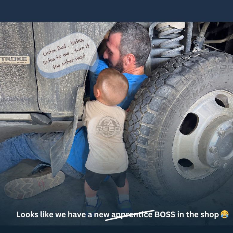 A mechanic with a young apprentice under a truck at Reed Automotive
